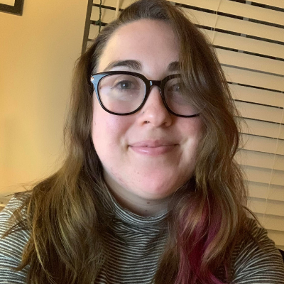 Danielle is smiling for a selfie. She's wearing dark-framed glasses and has a pop of pink in her brown hair. There is window blinds behind her.
