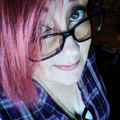 Jessica is smiling for a selfie. She's wearing dark-framed glasses and has pink hair. The background is dark behind her.