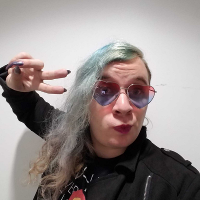 Light posing for a selfie. They have colorful hair and they're wearing pink heart glasses. They are making a peace sign with their hand. Background is white behind them.