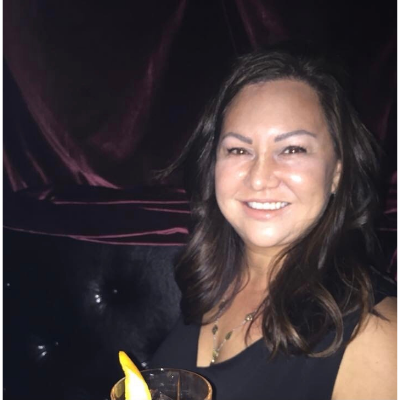 Stacey is smiling and facing a camera. She has dark brown hair. Background is dark behind her.
